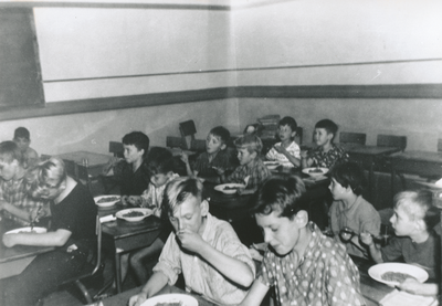 876378 Afbeelding van een groep kinderen die tussen de middag scholeneten krijgt, in een klaslokaal op de Nederlands ...